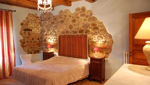 Un dormitorio con pared de piedra, vigas de madera en el techo, una cama con cabecero estampado, mesitas de noche con lámparas y una ventana con cortinas.