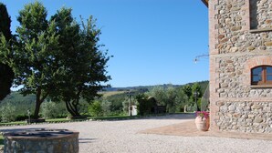 Un edificio de piedra con un camino de grava, un pozo de piedra y un árbol grande al fondo.