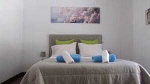 Un letto rifatto con precisione, con testiera grigia, cuscini bianchi e verdi, e asciugamani blu e bianchi.