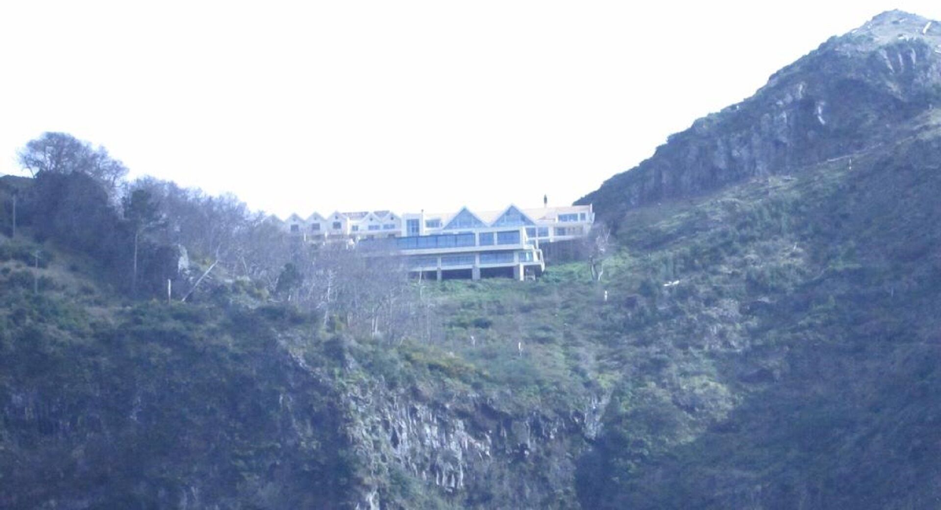 Un edificio de varios pisos con un estilo arquitectónico moderno, ubicado en una colina con vegetación densa.