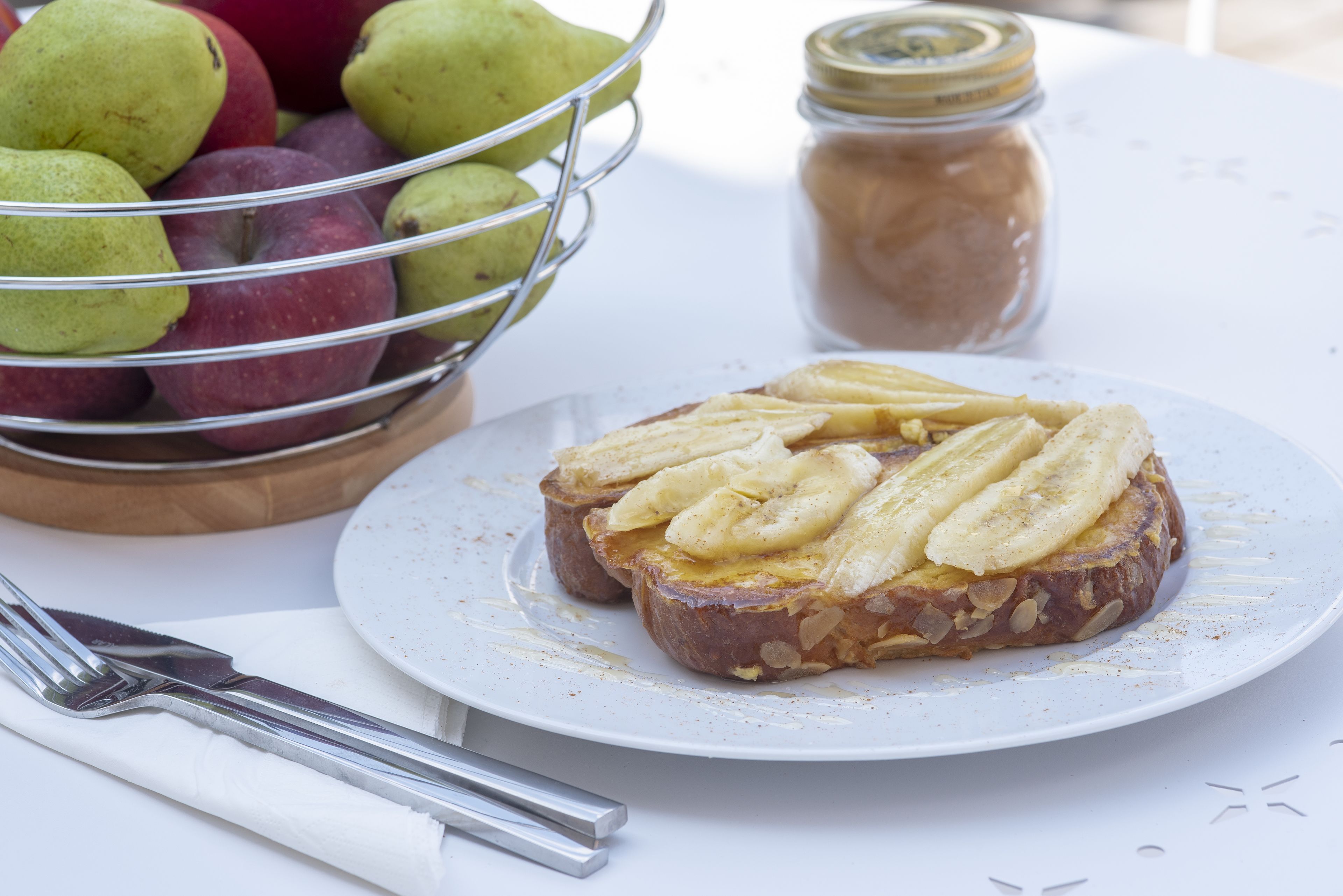 A plate with a sandwich topped with banana slices, a bowl of pears, and a jar of spread on a table.