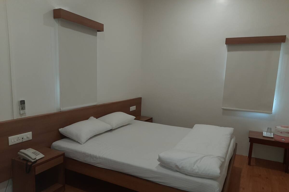 Een netjes opgemaakt bed met witte lakens, een houten hoofdbord, een nachtkastje met een telefoon en een klein nachtkastje met een glas en een doos.