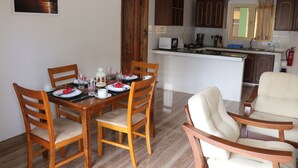 Un comedor con una mesa de madera dispuesta para cuatro personas, rodeada de sillas de madera. Se ve una cocina al fondo con armarios de madera y un refrigerador blanco.
