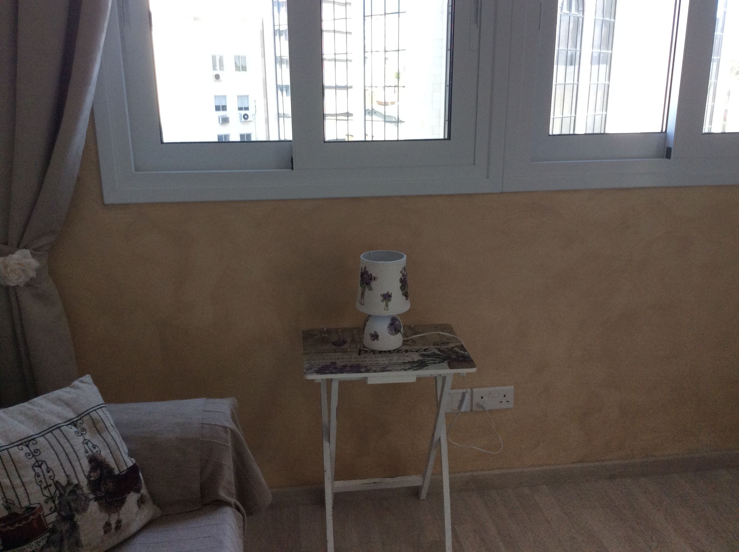 Ein Zimmer mit Fenster, einem kleinen Tisch mit Vase und einer Couch mit Zierkissen.