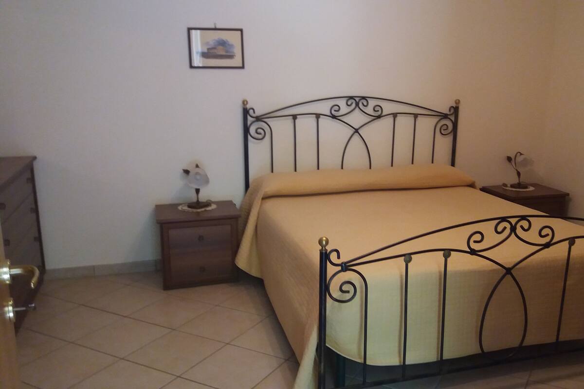 Un dormitorio con estructura metálica para la cama, una mesita de noche con lámpara y un aparador.