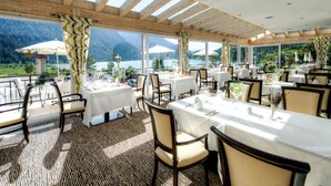 Un restaurante con mesas dispuestas para comer, amplios ventanales con vistas a las montañas y al lago, y una estructura de techo de madera.