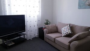 Un salon avec un téléviseur à écran plat posé sur un meuble, un canapé beige agrémenté de coussins à motifs, une petite table d’appoint avec une plante et une fenêtre ornée de rideaux légers.