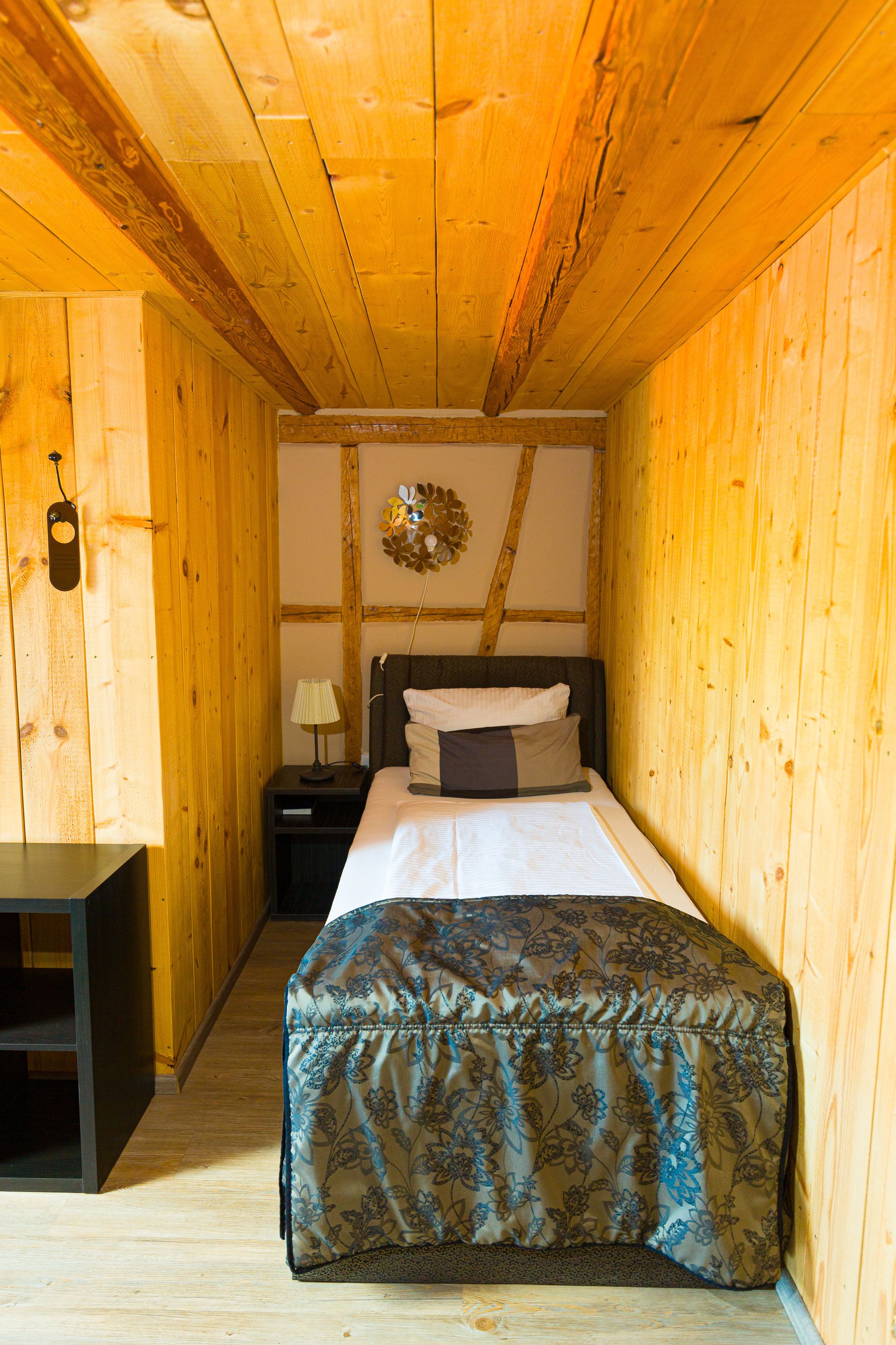 Ein schmaler Raum mit Holzboden, einem Bett, einem Nachttisch und einem kleinen Tisch.