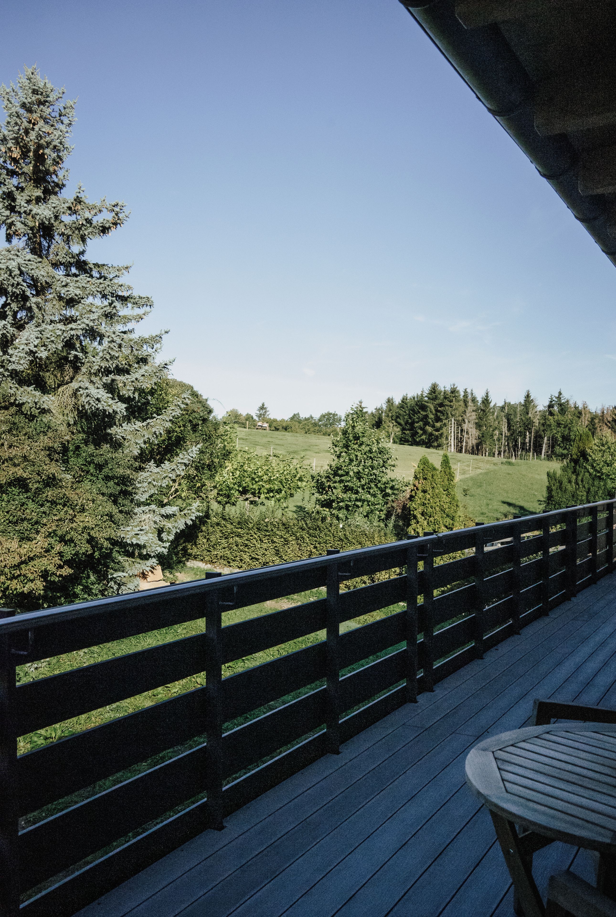 A wooden deck with a table and chairs, overlooking a green landscape with trees and a distant building.
