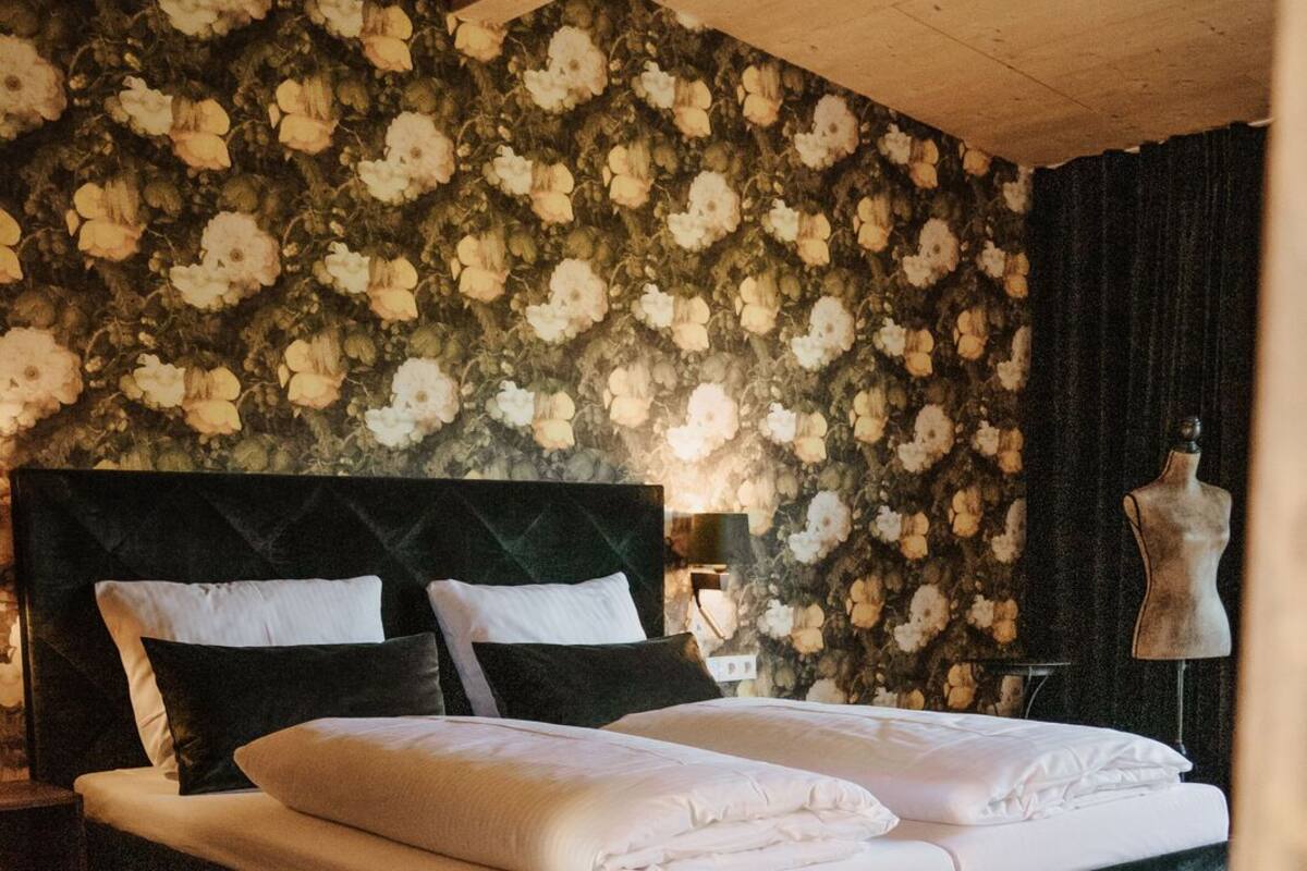Une chambre à coucher avec un grand lit, une tête de lit sombre, un papier peint à motifs floraux, un plancher en bois et un mannequin placé dans un coin.