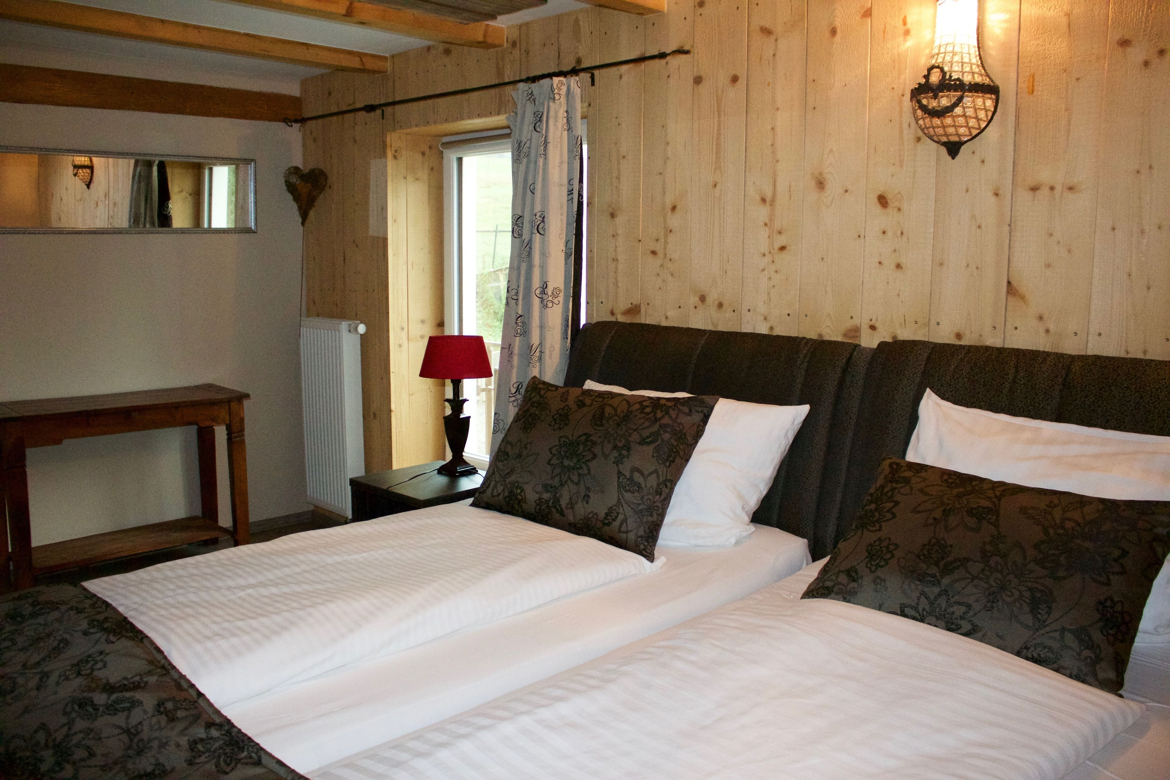 A double bed with white bedding and floral-patterned pillows, a wooden headboard, a small bedside table with a red lamp, and a mirror on the wall.