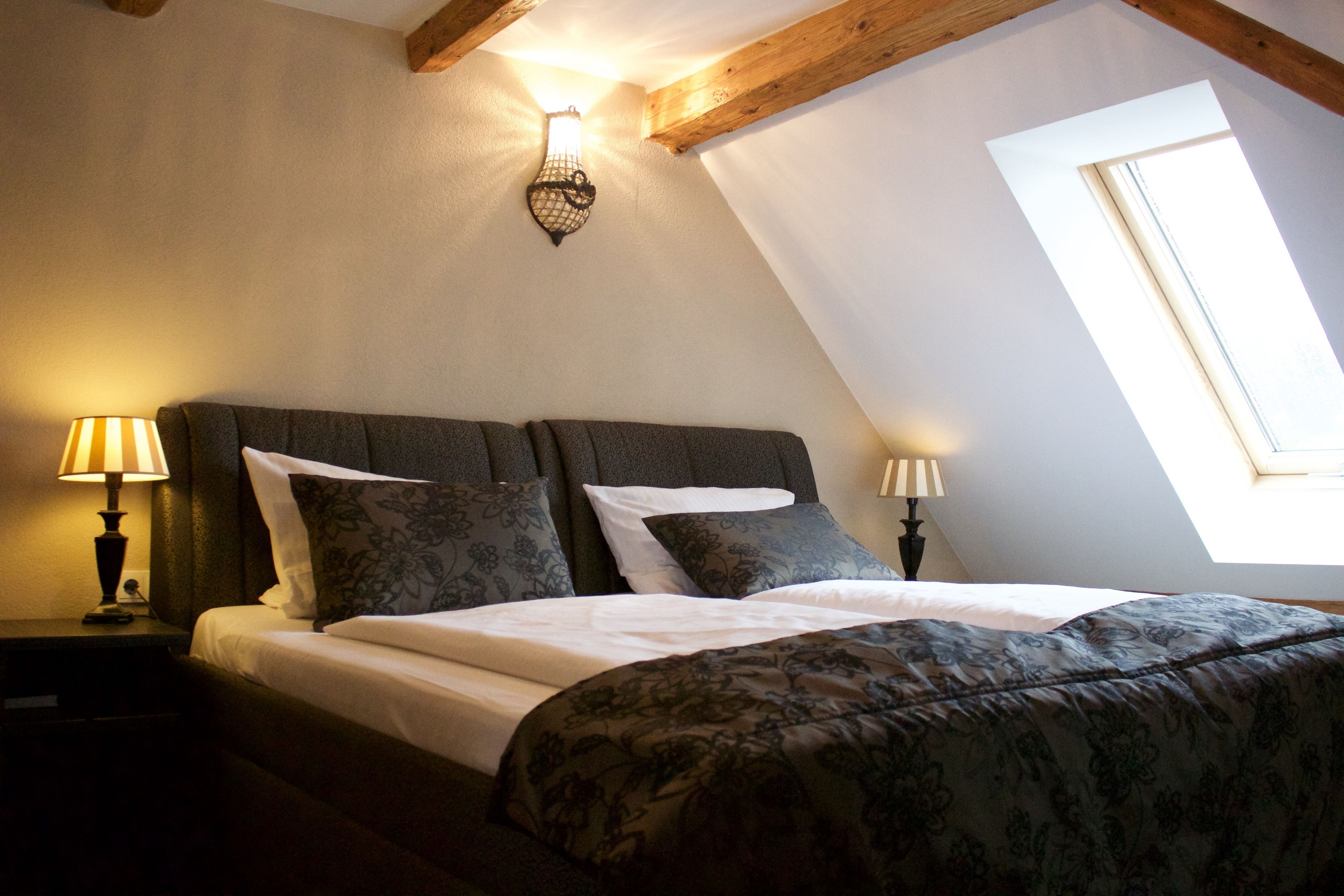 Ein Schlafzimmer mit schrägem Dach, zwei Nachttischlampen, einem Bett mit floralem Bettzeug und einem Fenster mit Tageslicht.