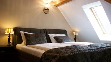 Ein Schlafzimmer mit schrägem Dach, zwei Nachttischlampen, einem Bett mit floralem Bettzeug und einem Fenster mit Tageslicht.