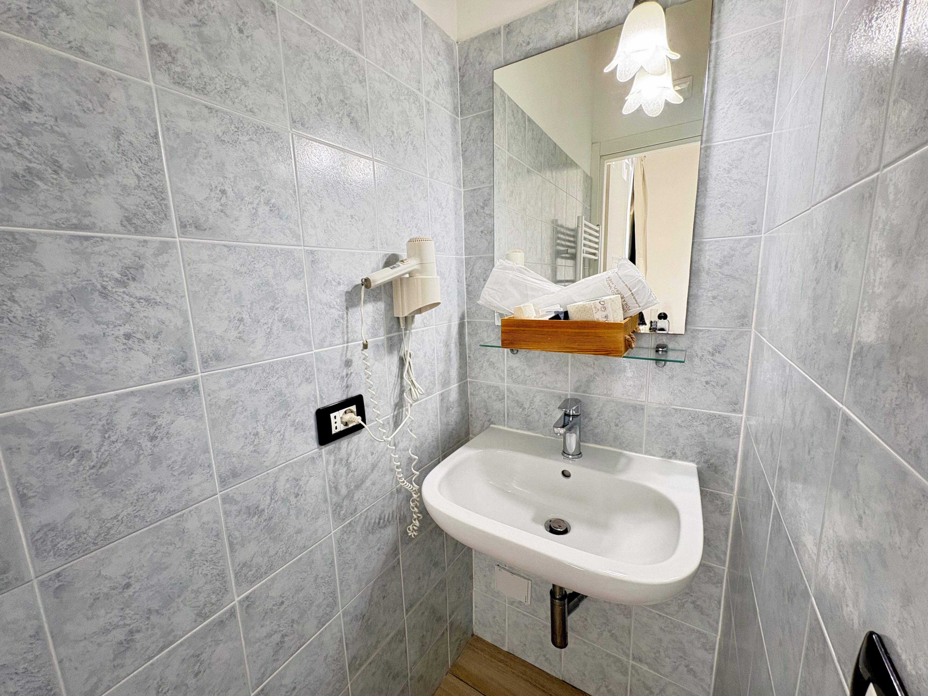Une salle de bain avec des carreaux de marbre gris, un lavabo blanc, un miroir et un sèche-cheveux fixé au mur.