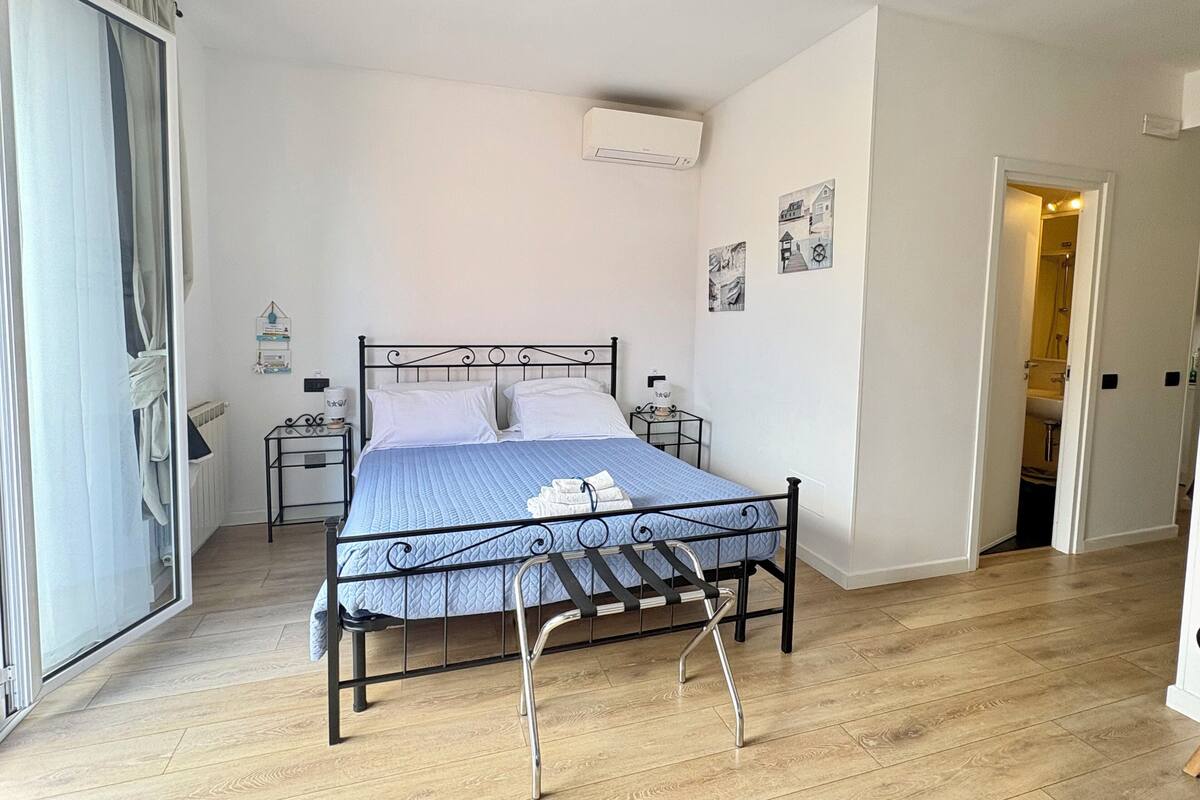 A bedroom with a metal bed frame, a blue quilted bedspread, a bedside table with a lamp, and a wall-mounted air conditioner.