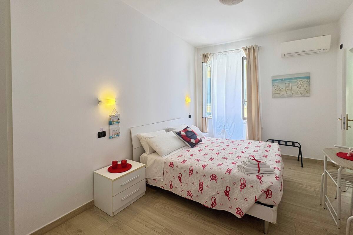 A bedroom with a bed, bedside table, wall-mounted lamp, window with curtains, and a small table with a chair.