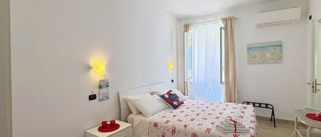 A bedroom with a bed, bedside table, wall-mounted lamp, window with curtains, and a small table with a chair.
