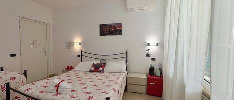 A hotel room with a bed, bedside tables, an air conditioning unit, a painting, and a window with curtains.