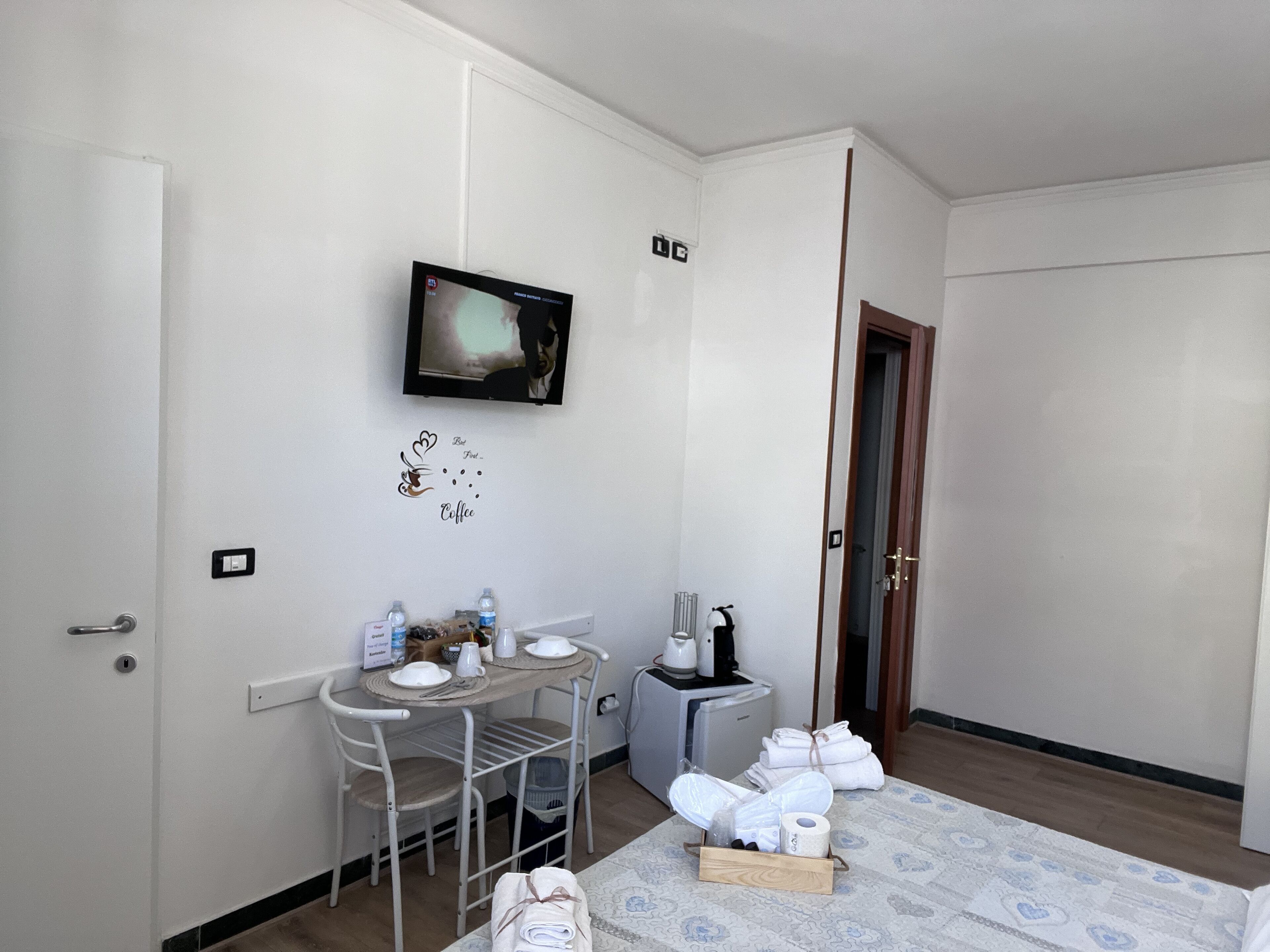 A small, clean room with a bed, a TV mounted on the wall, a small table with a chair, and a bedside table with toiletries.