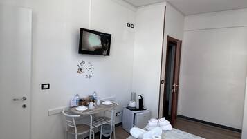 A small, clean room with a bed, a TV mounted on the wall, a small table with a chair, and a bedside table with toiletries.
