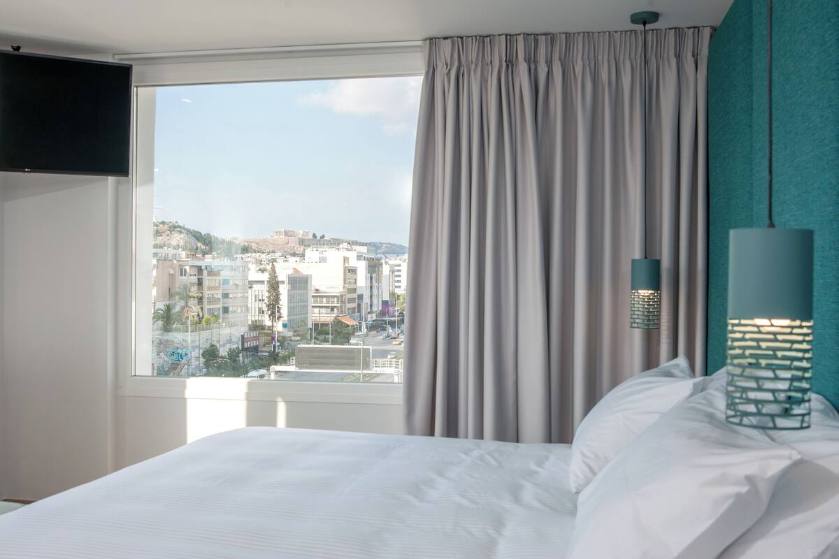 A hotel room with a bed, a TV mounted on the wall, a window with curtains, and a view of a cityscape.