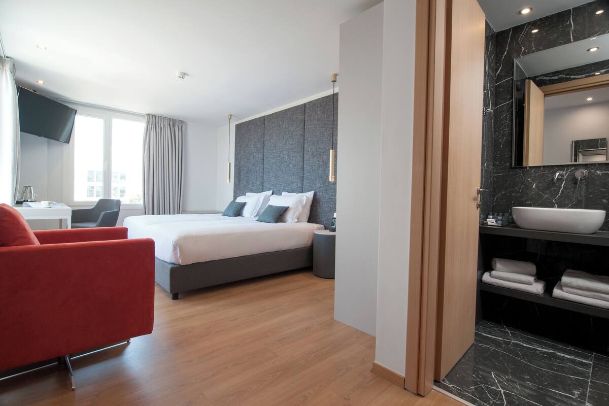 A modern hotel room with a large bed, a desk, a red armchair, and a bathroom with a large mirror and a white sink.