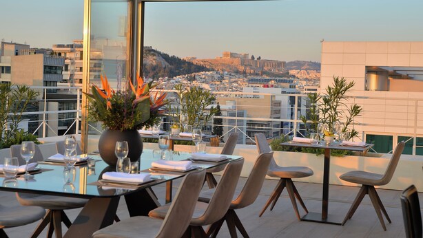 A rooftop dining area with a table set for two, overlooking a cityscape with historical buildings. - Athenaeum Smart Hotel (Athens)
