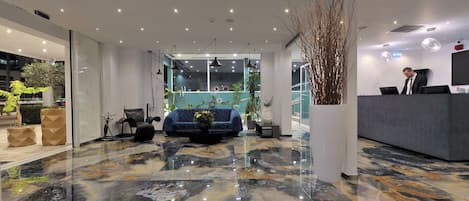 A modern hotel lobby with a marble floor, seating area, reception desk, and large windows.