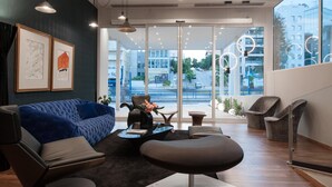 A modern living room with a blue sofa, black ottoman, and grey chairs. There are framed artworks on the wall, a coffee table with a vase, and a large window with a view of buildings and a street. - Athenaeum Smart Hotel (Athens)