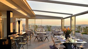 A rooftop restaurant with tables and chairs arranged for dining, large windows offering a city view, and a modern architectural design. - Athenaeum Smart Hotel (Athens)