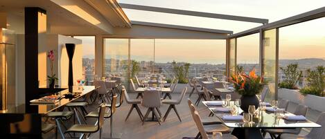 Un restaurante en la azotea con mesas y sillas dispuestas para comer, amplios ventanales que ofrecen vistas panorámicas de la ciudad y un diseño arquitectónico moderno.
