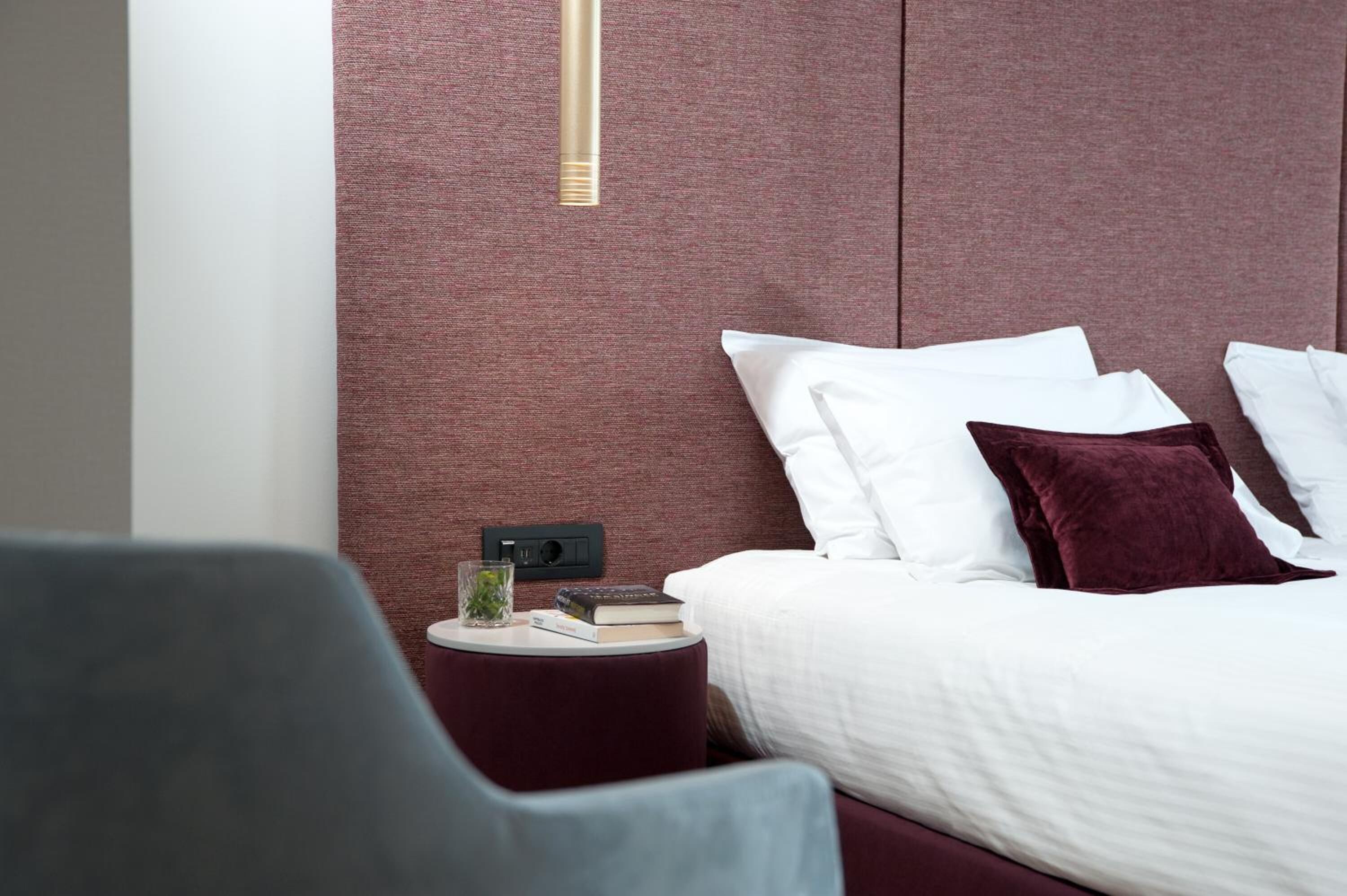 A modern hotel room with a large bed, a small side table, a chair, and a bedside table with a lamp.