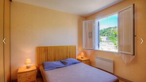 A bedroom with a wooden headboard, a bed with blue bedding, two bedside tables with lamps, and a window with a view of greenery and buildings. - Gîte Calzolari in Corsica (Castifao)