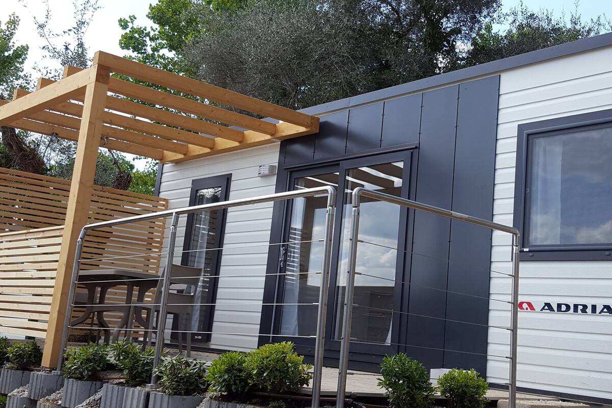 Een moderne stacaravan met een pergola, een schuifdeur en een raam met het merk 'ADRIA' erop.