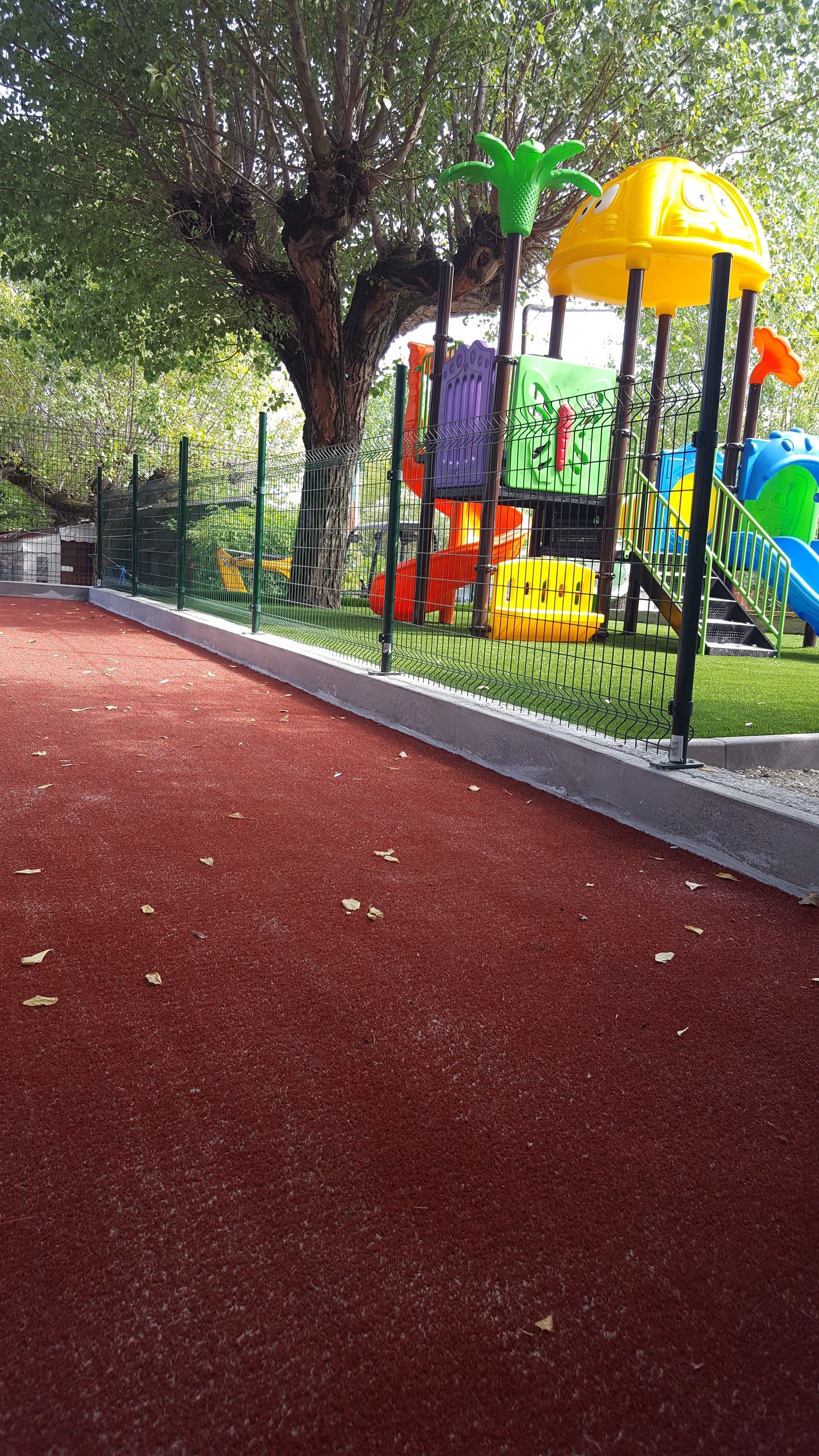 A playground with a red rubber surface, surrounded by a green fence, and colorful play structures.