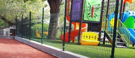 A playground with a red rubber surface, surrounded by a green fence, and colorful play structures.
