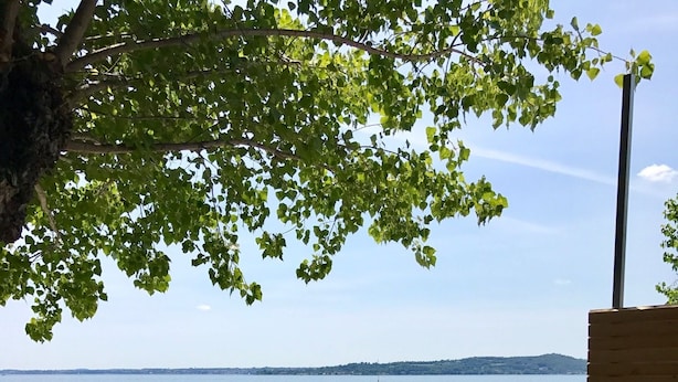 Une terrasse en bois, agrémentée de chaises et de tables en métal, offre une vue sur une mer calme où voguent des bateaux.