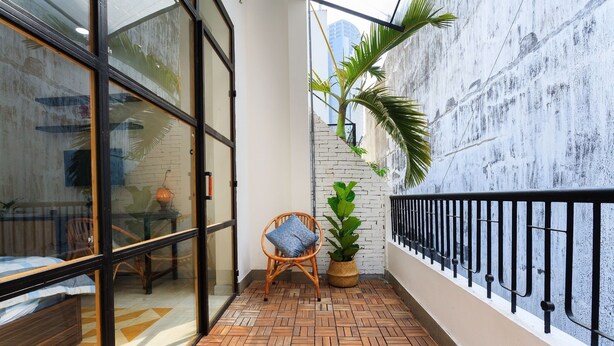 Un balcón con suelo de patrón espiga, una silla de mimbre con cojín, una planta en maceta y vistas de un paisaje urbano a través de un ventanal.