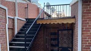Un étroit couloir extérieur avec un escalier en métal menant à une porte en bois ornée de panneaux décoratifs.