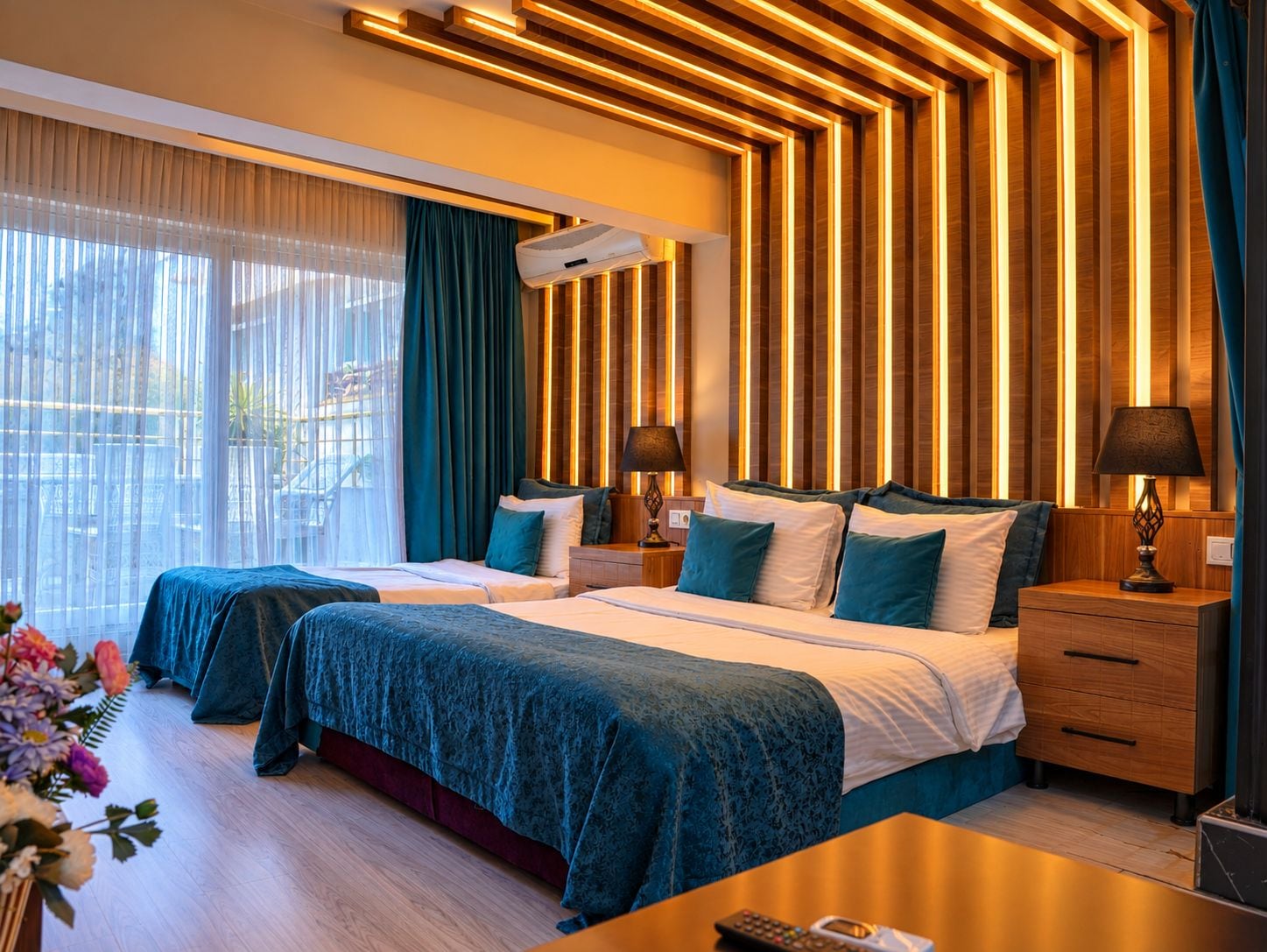 A hotel room with two beds, a wooden ceiling with vertical lighting, a large window with curtains, and a bedside table with a lamp.