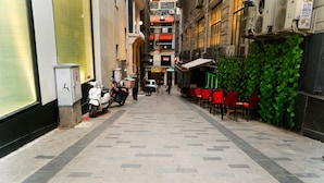 Une ruelle étroite, avec un scooter garé sur le côté gauche, une rangée de chaises rouges sur le côté droit, et un bâtiment doté d’un mur vert et d’une enseigne.