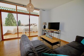 Un salon moderne avec un canapé, un téléviseur posé sur un meuble et une vue sur une terrasse extérieure, accessible par une porte vitrée.