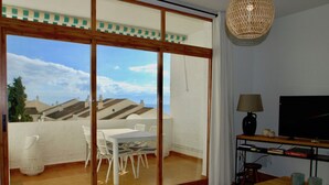 Ein Wohnzimmer mit Fernseher, Tisch und Sesseln, ein Balkon mit Tisch und Stühlen sowie ein großes Fenster mit Blick auf Häuser.