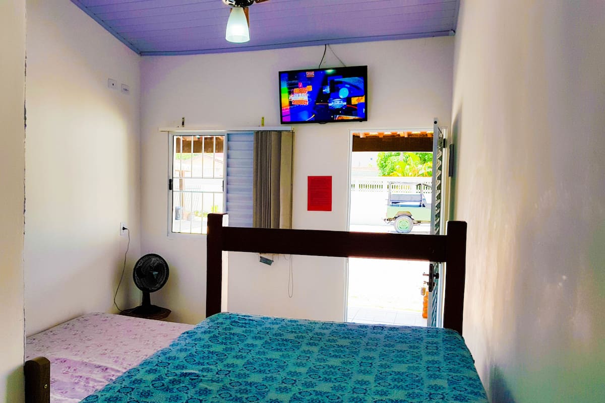 A bedroom with a bed, a ceiling fan, a TV on the wall, and a window with a view of the outside.