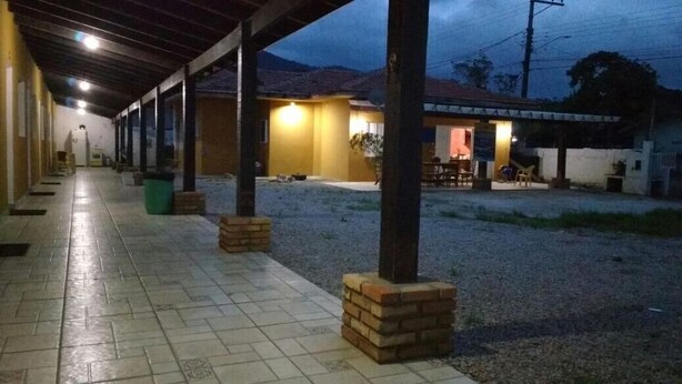 Un espacio al aire libre cubierto, con suelo de baldosas, pilares de ladrillo y un edificio al fondo.