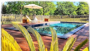 A swimming pool with a wooden deck, surrounded by a wooden fence and palm trees. - Au Clair Des Landes (Castets)