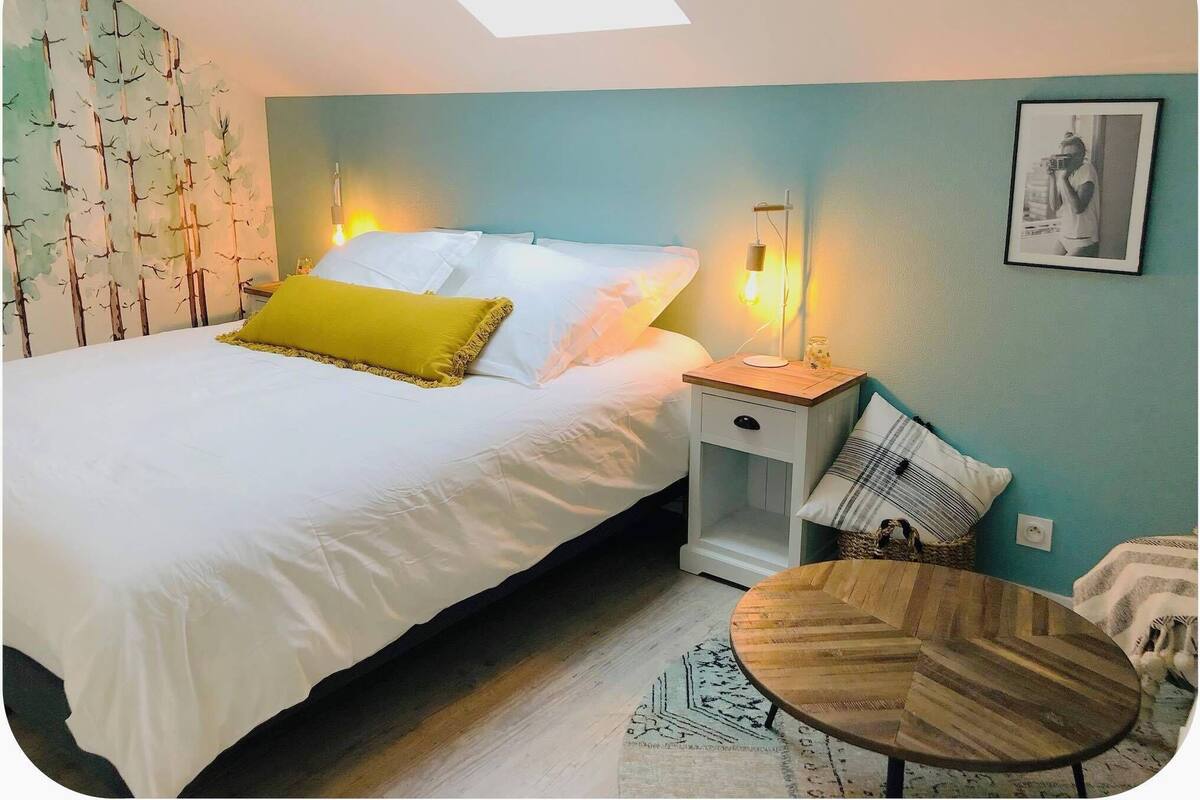 A bedroom with a bed, bedside tables, a round wooden table, and a wall with a framed picture.