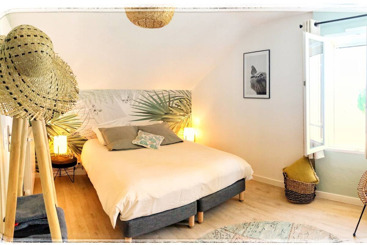 A bedroom with a bed, a nightstand, a lamp, a woven chair, a picture on the wall, and a window.