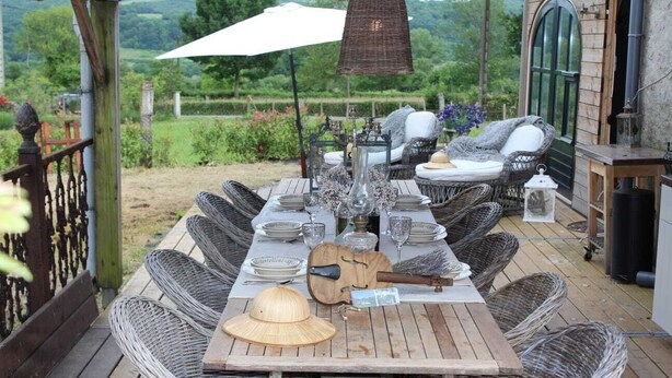 An outdoor dining area with wicker furniture, a wooden table set with plates, glasses, and a hat.