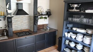 A kitchen with dark cabinetry, a wooden countertop, a sink, and a shelf with white dishes and glasses.